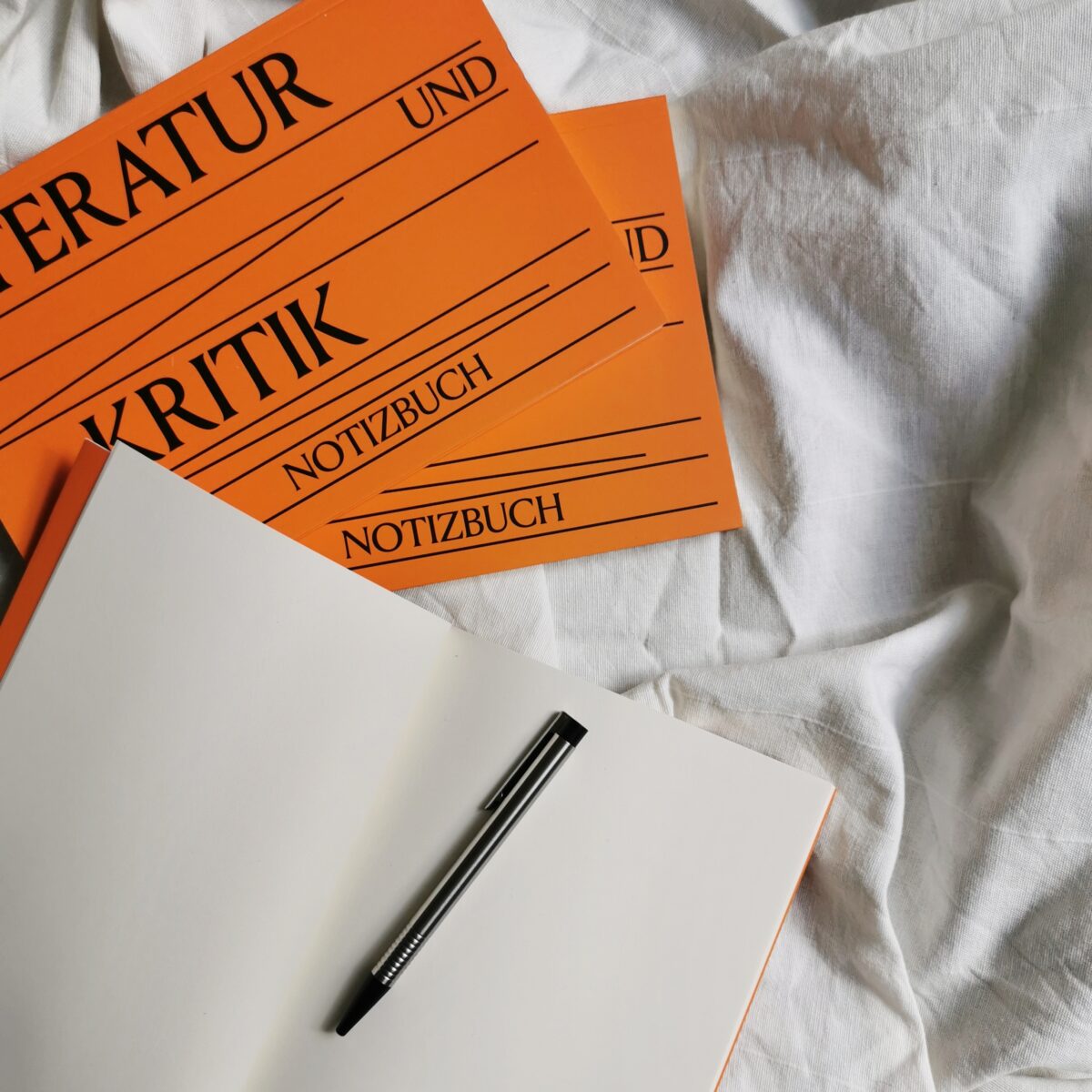 offenes Notizbuch mit Stift, zwei geschlossene Notizbücher in orange auf einer weißen Decke
Auf den Notizbüchern steht "Literatur und Kritik"